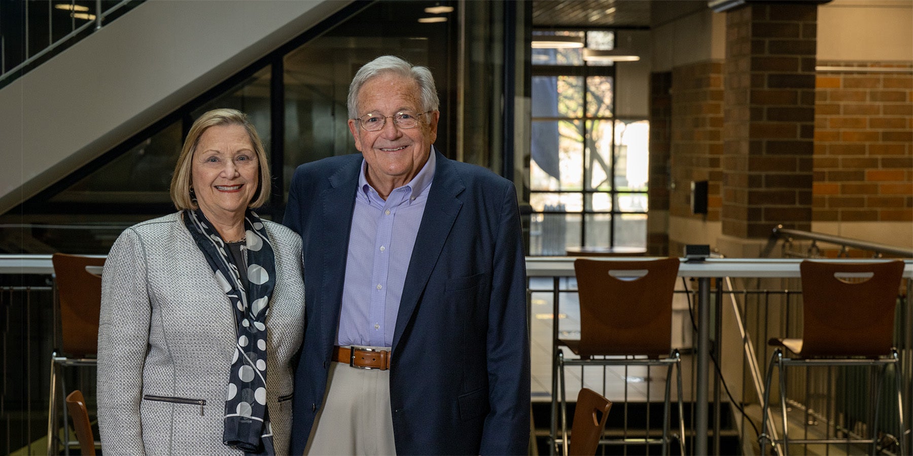 Gloria (left) and Steve (right) Bailey stand smiling in the Bailey College of Engineering & Technology Building. She is wearing a beige business casual suit with a black scarf that has large white circles on it. He is wearing a dark blue sport coat with light blue shirt, beige khakis, and brown belt. The visible interior of the building is dark, with outdoor sunlight visible through windows in the back of the building.