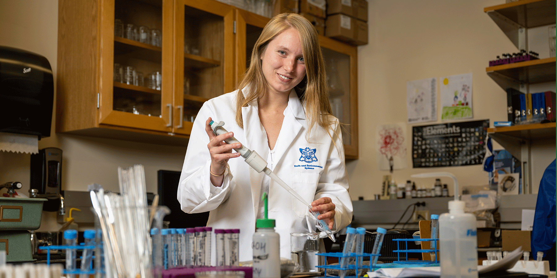 A female student with straight blonde hair stands in a laboratory room, wearing a white lab coat. She holds a long syringe. Beakers are on the table. 