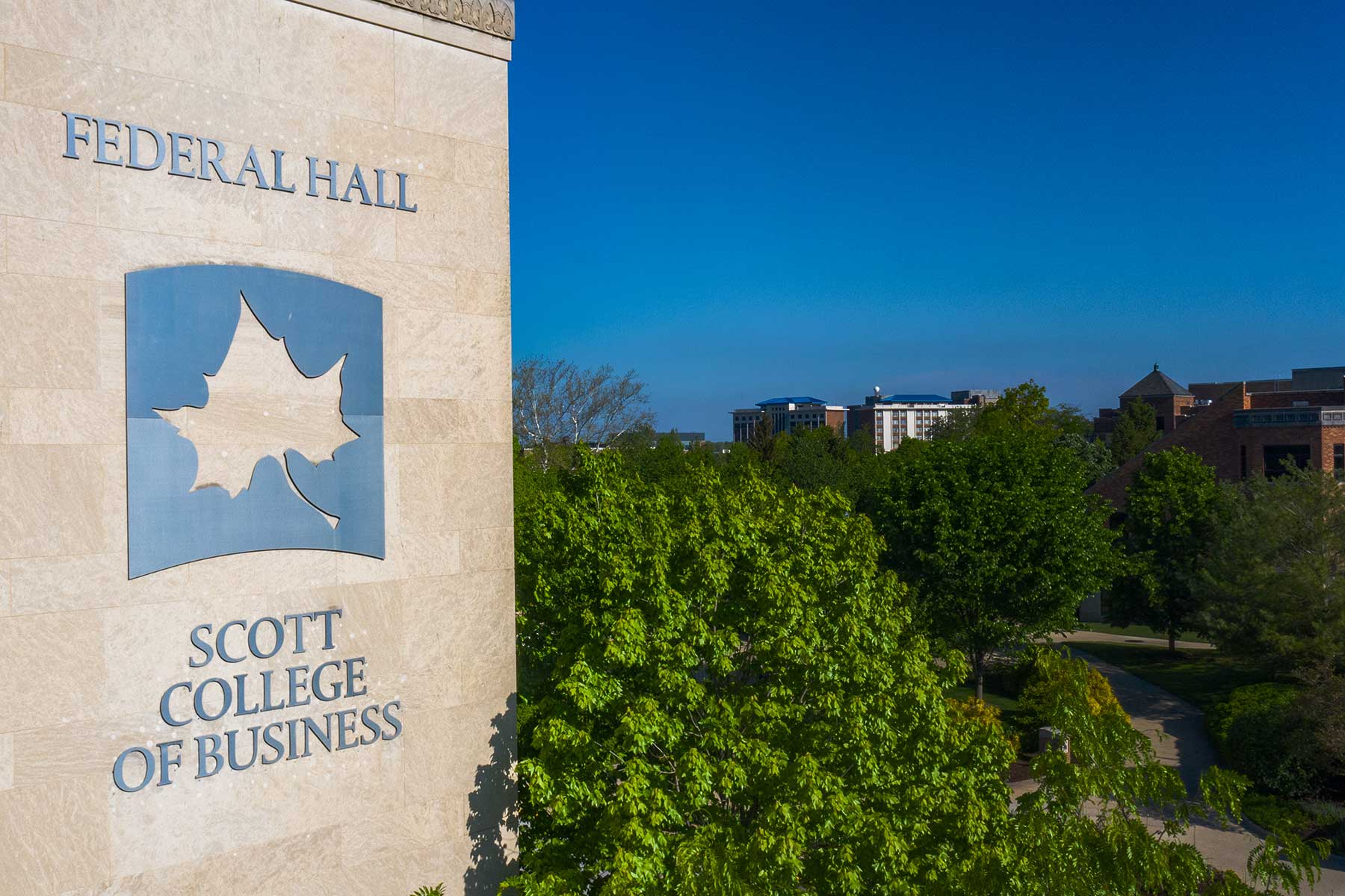 The grey sandstone of Federal Hall is visible on the left, with the building’s name in blue, and beneath that the grey Sycamore leaf cutout on a blue background, followed by ‘Scott College of Business.’ To the right of the building, green trees are visible, with the tops of other buildings visible in the background.