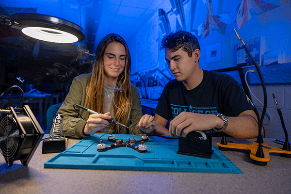 Two students are working in a blue-lit laboratory space. On the left is a white female student with long straight brown hair. She wears a dark green jacket with a grey undershirt. On the right is a white male student with short brown hair. He wears a black T-shirt with blue lettering on the front. The woman holds a soldering iron. Technology equipment is visible on the table in front of them with a blue tray under the equipment. A lamp light sits left of the equipment.
