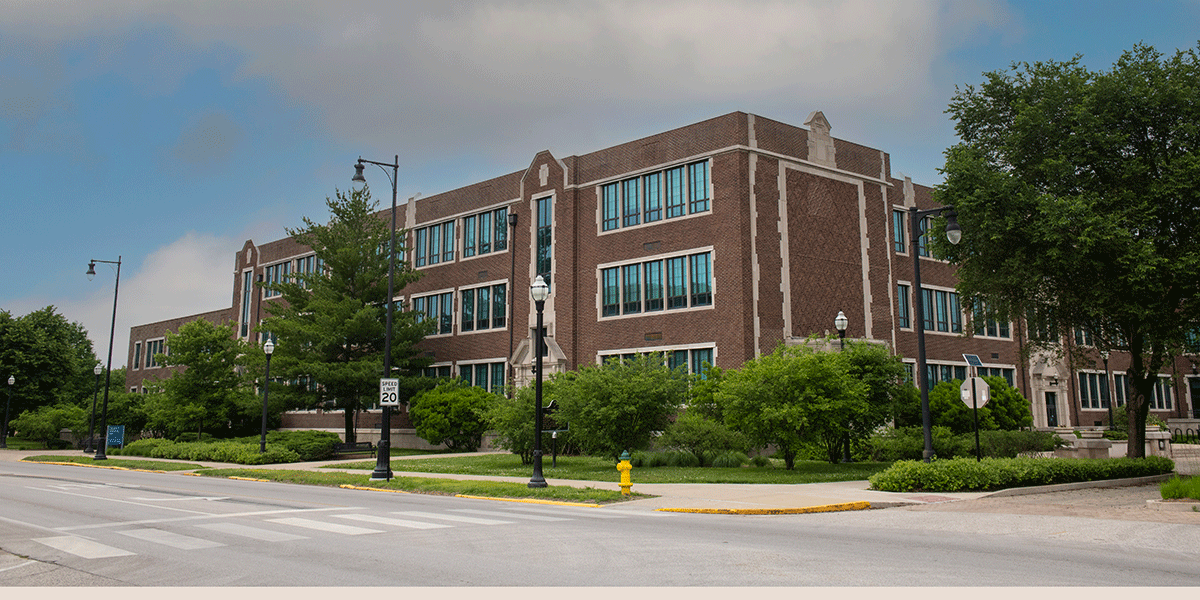 A red brick building with white trim, as viewed from the street. A lamppost, trees and greenery, and a speed limit sign showing 20 mph are all visible in front of the building. Blue sky and greyish-white clouds are visible above.