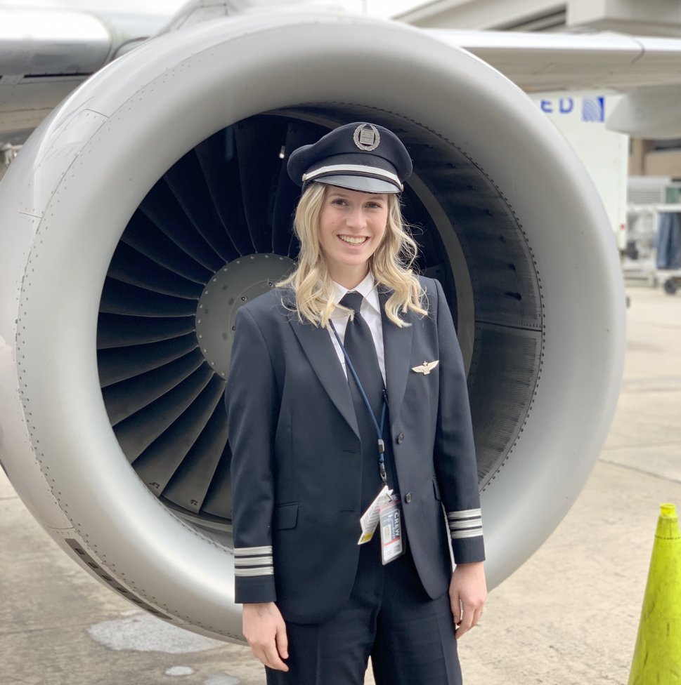 A smiling young blonde woman in a black and white pilot’s uniform and cap stands in front of an airplane’s silver-and-gray turbine engine.  