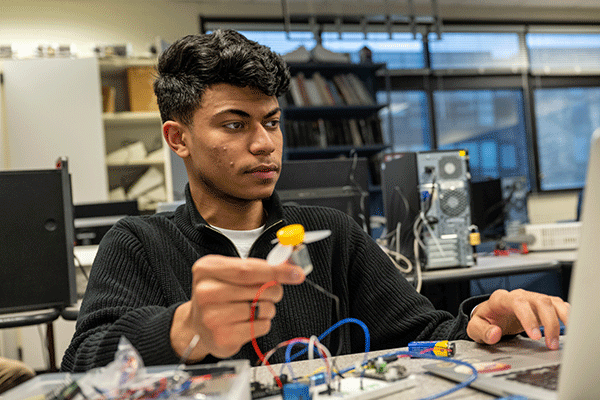 A male student with a dak olive complexion and short black hair sits at a table with a laptop in front of him. He wears a black zipped-up jacket with a white undershirt. Blue and red wiring is visible on the table. He holds a small grey electrical device in his right hand, with a yellow top and grey propellers. Other electrical devices are visible on the table. Computer monitors, a bookcase with stacks of books, and a row of windows are visible in the background. 