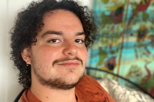 Close-up of a student with curly brown hair with a matching beard and moustache. The student is staring at the camera and wears an orange shirt. In the background is a blue, red, and yellow floral art piece.