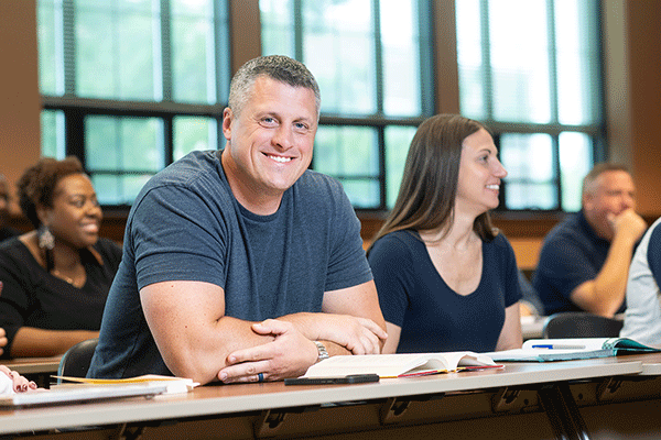 A middle-aged white male with short greying hair and a grey t-shirt sitting at a table in a classroom looks toward the camera and smiles with a diverse group of classmates in the background.  