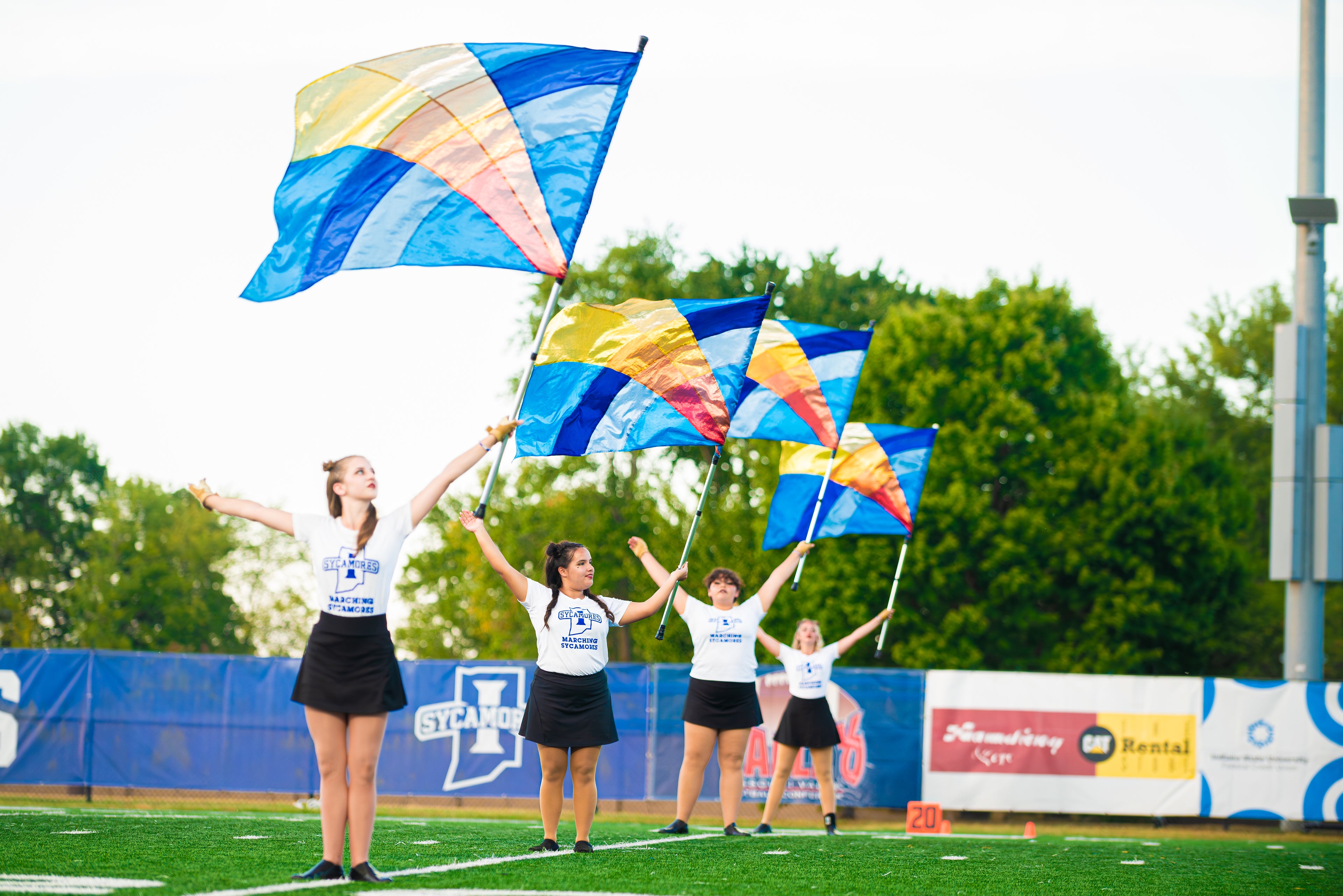 A group of people on a sports field wave large blue and yellow flags. They wear matching shirts and black skirts, performing in a line formation with trees in the background.