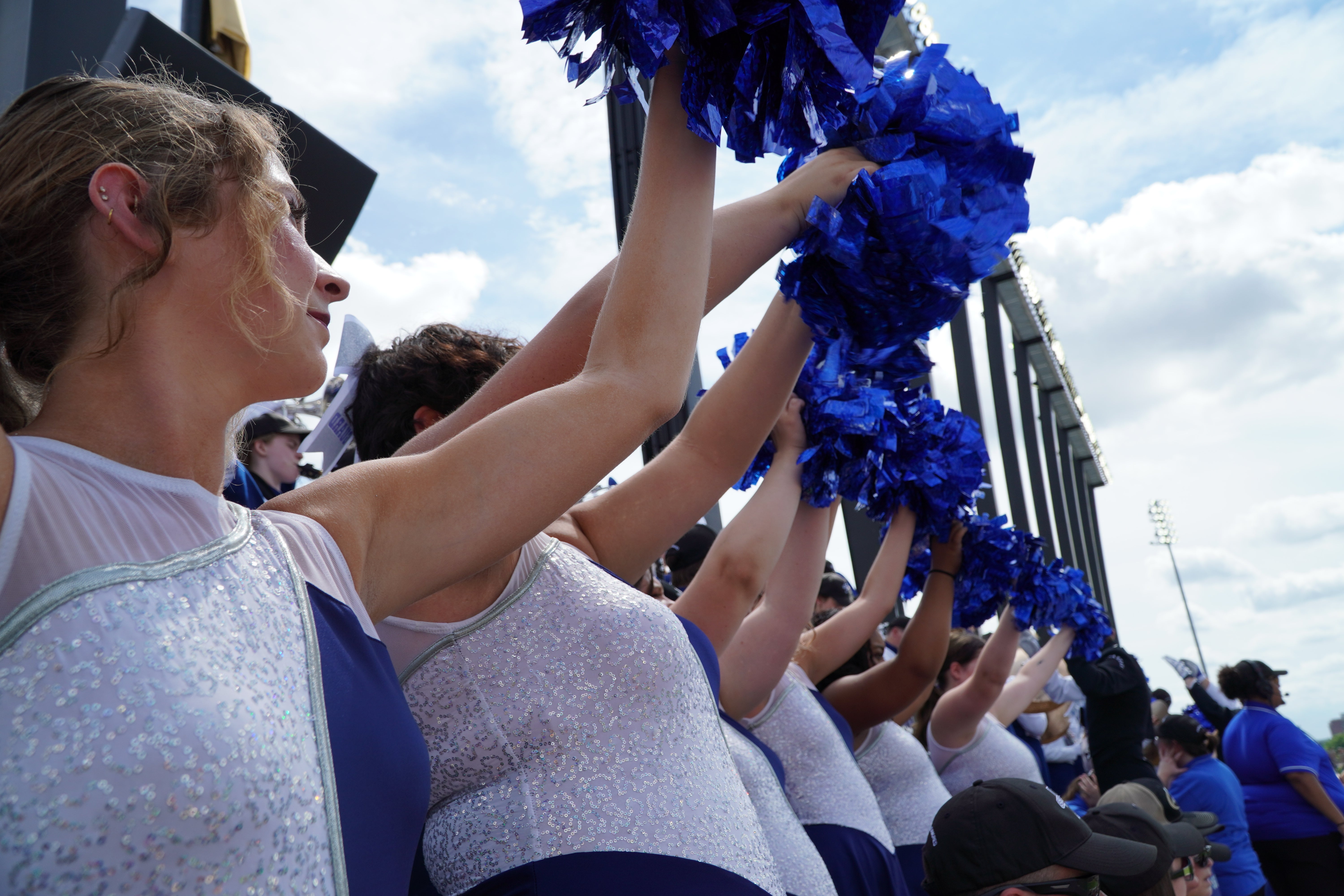 Cheerleaders in blue and white uniforms raise blue pom-poms in the air during an event, with a bright sky in the background.