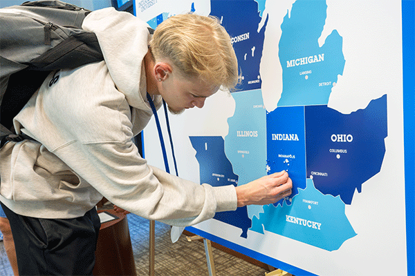 A white male student with short blonde hair stands before a display with a picture of Midwestern states in various blue shades. He wears a white hoodie, black pants, and a black backpack. The map has tiny blue pins and the student is placing a pin towards the middle of Indiana.