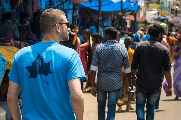 A white male with short brown hair wearing sunglasses and a blue T-shirt with a black leaf on the back is walking through a busy market street. The market is crowded with people, some wearing colorful clothing. Shops line the street, covered with blue tarps and various goods on display.