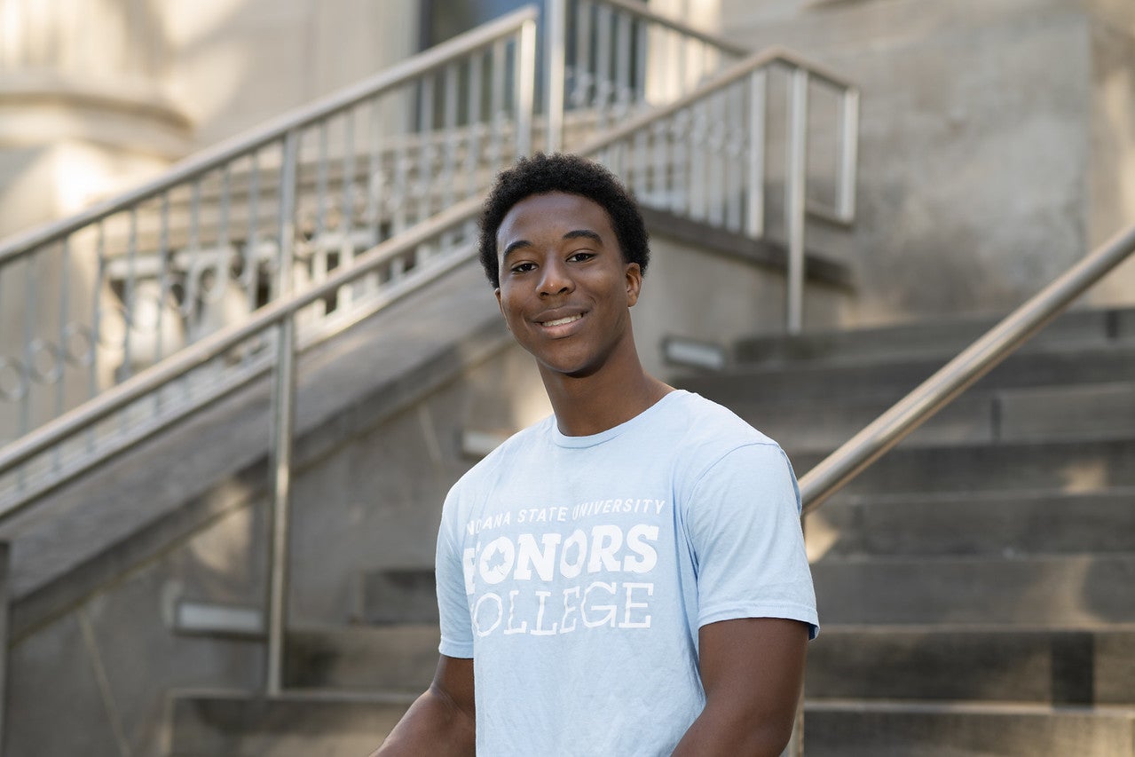 A Black male stands in front of a staircase. He is wearing a light blue T-shirt that reads "Indiana State University Honors College." The background includes part of a building and metal railings along the stairs.