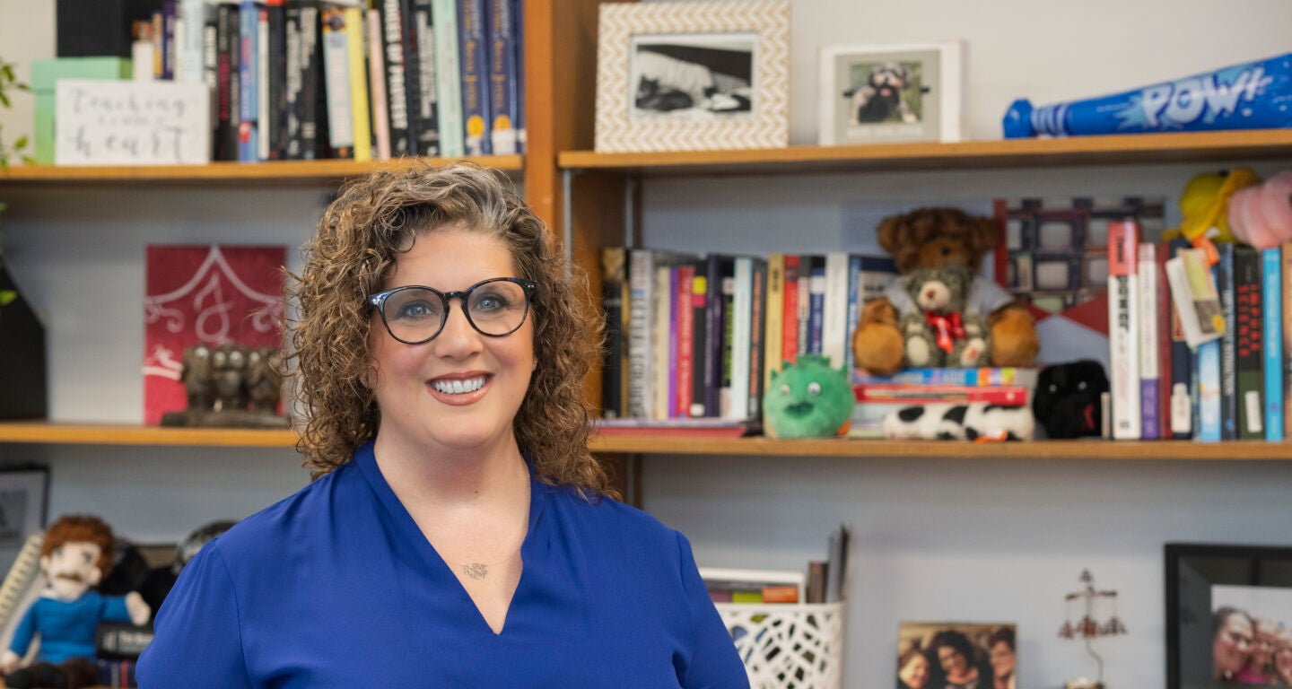 A white female with curly brown hair is wearing a blue shirt and standing in front of a bookshelf filled with various books, framed photos, and stuffed animals. The bookshelf has multiple shelves containing colorful books, a green plush toy, a teddy bear, and other decorative items.