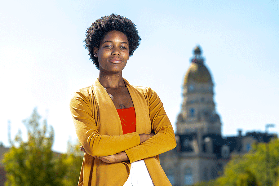 A Black female stands confidently with her arms crossed, wearing a yellow jacket over an orange top. The background features a historic building with a dome and trees.