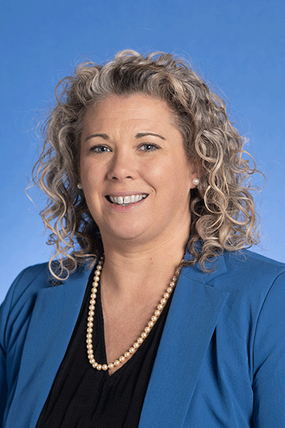 A smiling woman with curly, gray-blonde hair wears a blue blazer, black top, and pearl necklace, She stands against a solid blue background.