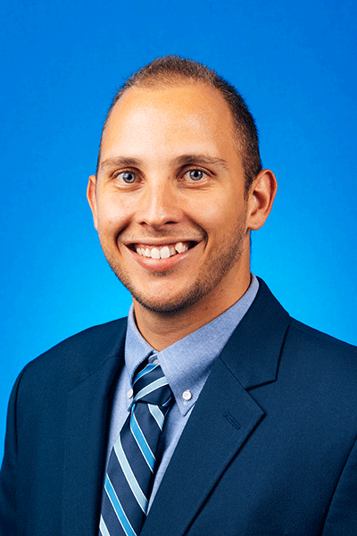 A smiling man in a dark blue suit jacket, light blue-striped dress shirt, and blue-striped tie, stands against a solid bright blue background.