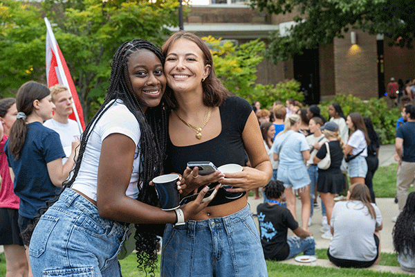 Two women are captured in a photo, posing on the grass with smiles, surrounded by greenery.  