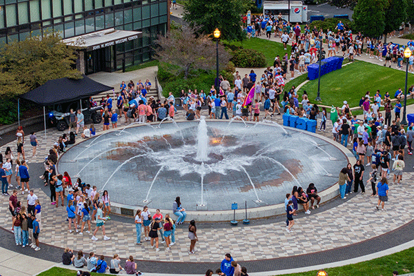 Many people are assembled around the Indiana State fountain, creating a vibrant scene in a bustling outdoor area.  
