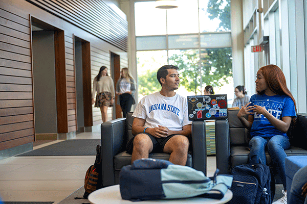 Two students in a lobby, sitting in chairs and interacting with each other.