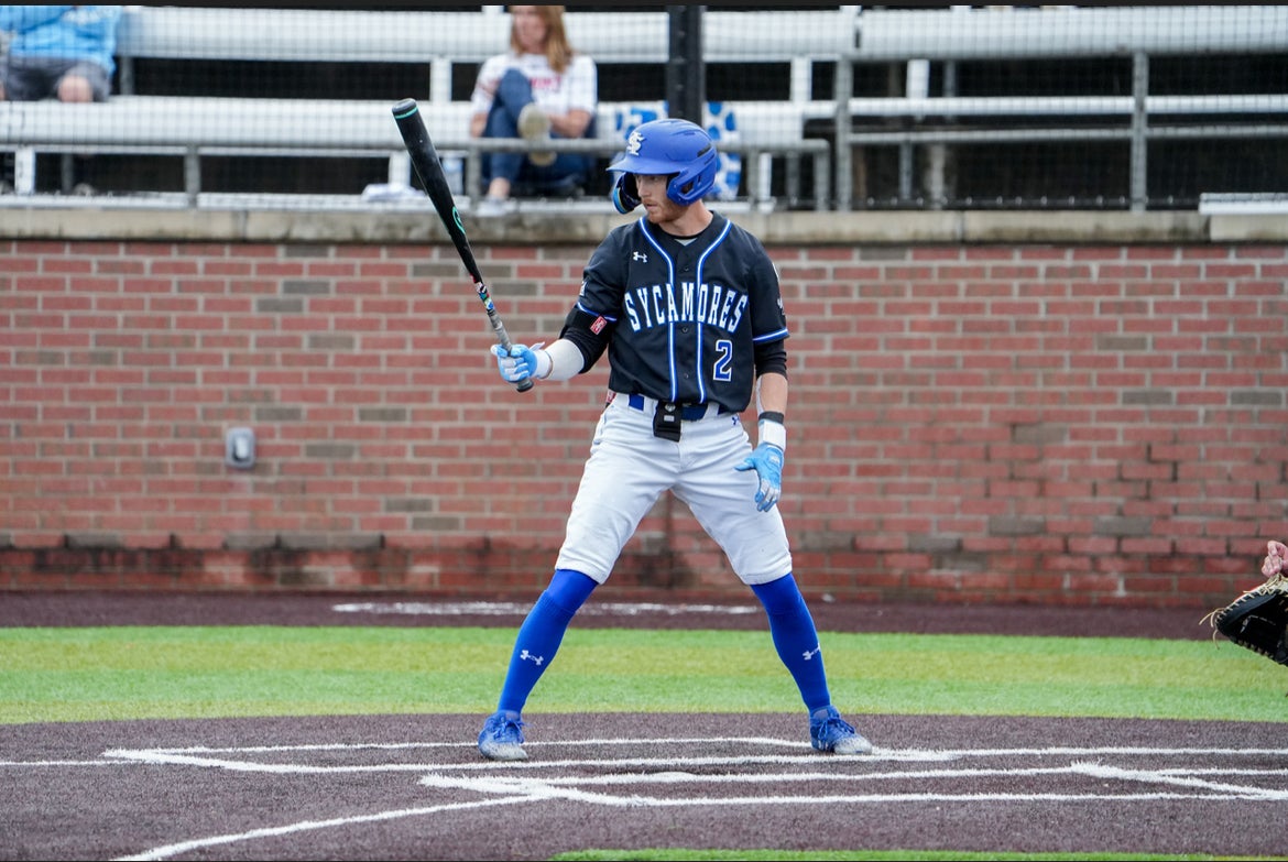 A baseball player in a blue helmet, black “Sycamores” jersey with the number 2, white pants, and blue socks stands in the batter’s box holding a bat. A catcher’s glove is visible on the right, with a brick wall in the background.