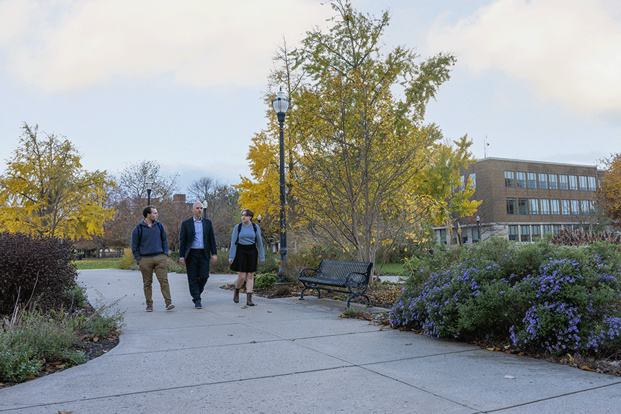Three people walk along a paved pathway in an outdoor campus setting with autumn foliage. Trees with yellow leaves, bushes, and flowers line the path. A bench and a lamppost are visible, with a multi-story building featuring large windows in the background.