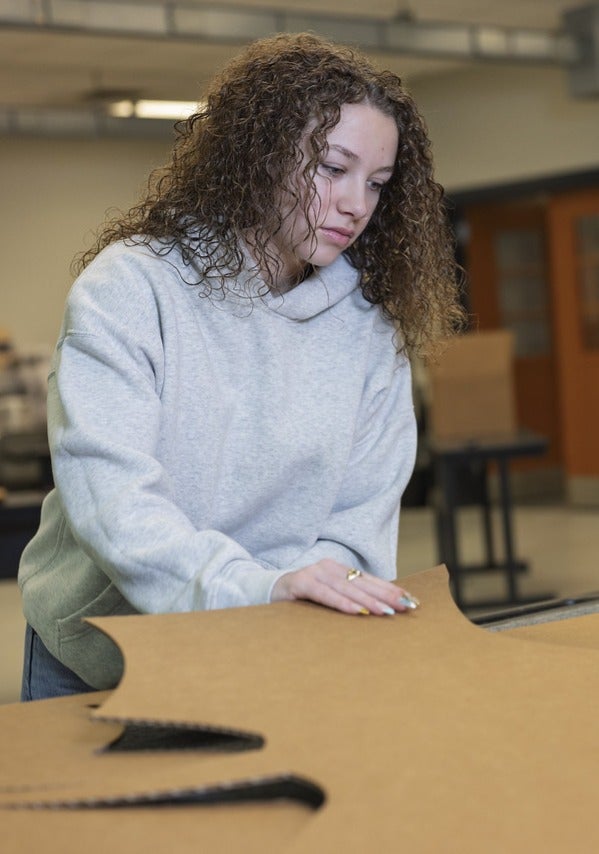 "A female person with curly brown hair wearing a light gray hoodie is working on a project involving large pieces of brown cardboard with intricate cut-out patterns. The individual is focused on cutting or shaping the cardboard in an indoor workspace that includes industrial equipment, a table, and machinery.