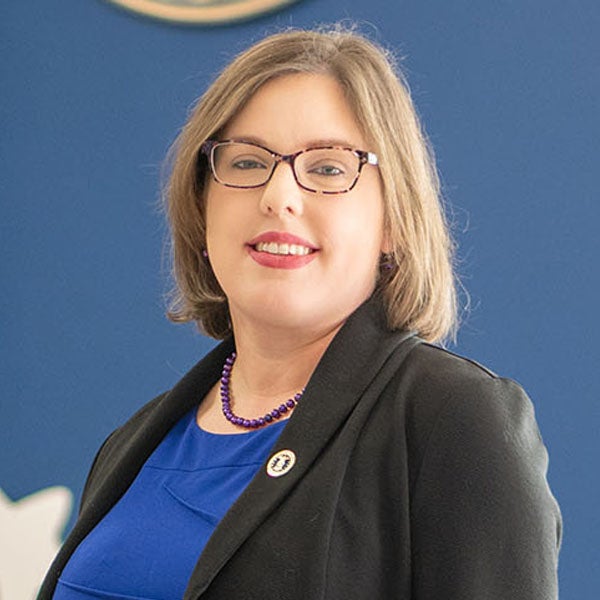 A smiling woman with shoulder-length, blondish-brown hair stands in front of a blue background. She wears glasses and a black blazer over a blue shirt, with a purple beaded necklace and a military service pin on the blazer's lapel.  