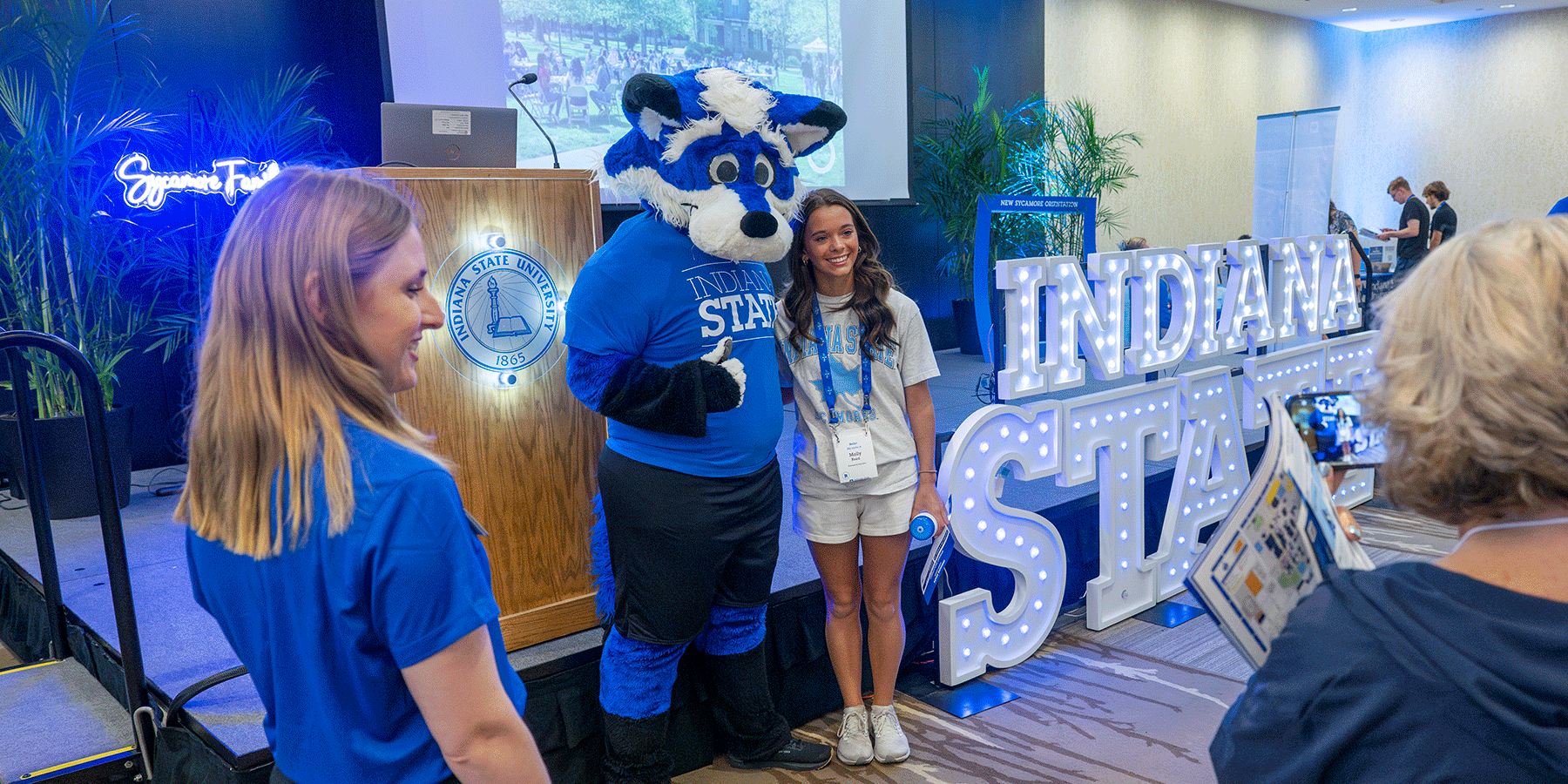 Indiana State mascot Sycamore Sam poses with a student at an indoor event. Letters spelling ‘INDIANA STATE’ are illuminated in the background.