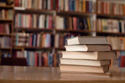 A stack of six books sits on a table in the library. The spines are facing away and cannot be read. In the background, shelves of colorful books are visible.