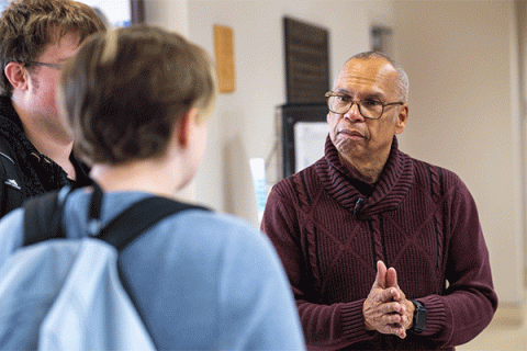 An older Black man with short, greying hair, and wearing glasses and a maroon sweater, is having a conversation with two young adults in an indoor space. One of them, with short brown hair and wearing a light blue top, is seen from the back with a sky-blue backpack on. The other is partly visible and has brown hair and glasses. The man's hands are pressed together as if emphasizing a point of discussion. 