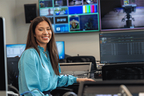 Student in a bright blue blouse seated in a media production lab, surrounded by monitors and video editing equipment, with a wall of broadcast screens in the background. 