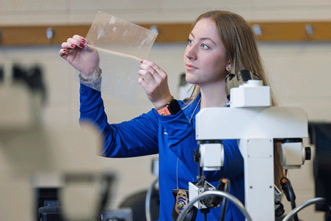 A young woman with straight blonde hair, wearing a blue jacket and a police badge, and with a focused expression on her face, examines a transparent paper while standing in a lab. A microscope and other equipment are visible in the foreground.