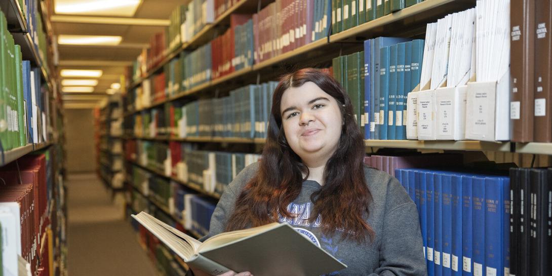 A white female student with long dark brown hair is standing in a library aisle, holding an open book. She wears a grey sweatshirt. Bookshelves are on both sides and are filled with various books, organized by color and size. The library is lit with fluorescent lighting. 